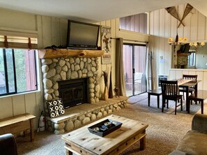 A cozy living room with a stone fireplace, a television mounted above it, and a dining area with a table and chairs. - ❤Comfy 2BD/2BA Condo❤WALK TO LIFTS👉Near Canyon Lodge❄Slps 6🐶Pets OK (Mammoth Lakes)