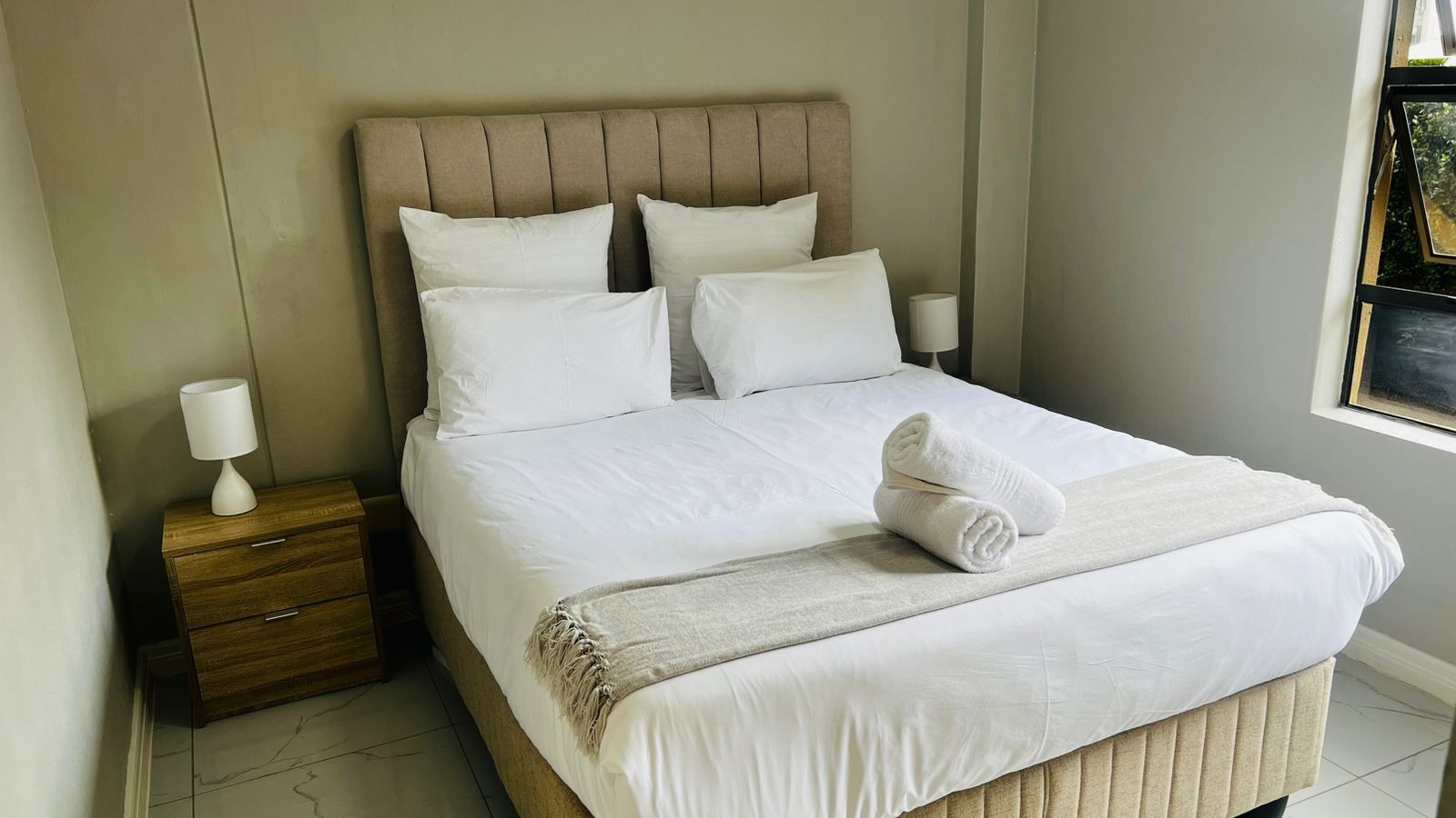 Een net opgemaakt bed met witte lakens, een beige hoofdbord en een nachtkastje met een lamp.