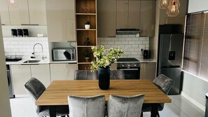A modern kitchen with a wooden dining table, grey upholstered chairs, a black vase with flowers, and built-in appliances. - Sandton Westpoint Luxury Apartments (Sandton)