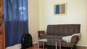En stue med en brun sofa, et glasbord, et træsidebord, en ventilator og en blå gardin.