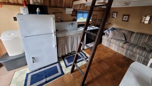 A compact kitchen with a refrigerator, stove, and sink. There is a wooden ceiling and a couch in the living area. - Blakley Hobbit Small Cabin (Viola)