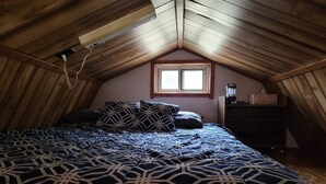 A bedroom with a sloped ceiling, a bed with patterned bedding, a window, and a dresser with a lamp. - Blakley Hobbit Small Cabin (Viola)