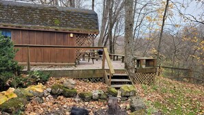 A rustic cabin with a wooden deck, a small wooden structure, and a fire pit area surrounded by rocks and fallen leaves. - Blakley Hobbit Small Cabin (Viola)