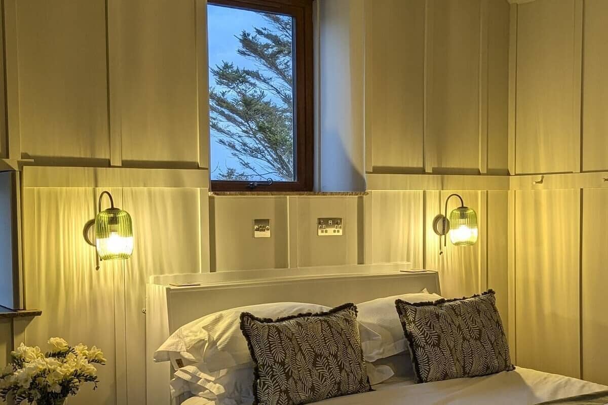 A bedroom with a bed, two wall-mounted lamps, a window with a view of trees, and a small table with a vase of flowers.