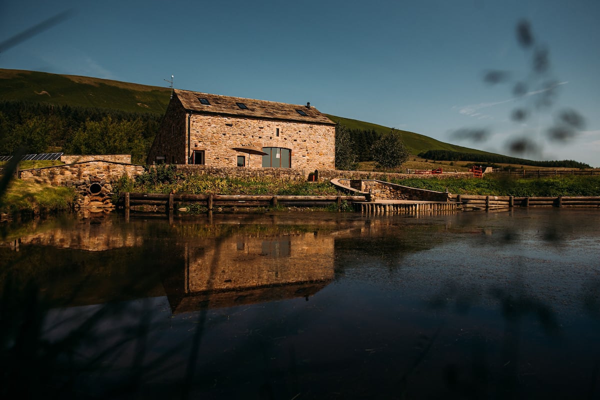 A stone building with a small door and windows, surrounded by a wooden fence and a body of water reflecting the structure.