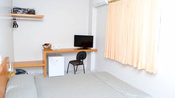 Una habitación con una cama, un escritorio con una computadora, una silla, un refrigerador pequeño y un estante con una bolsa.
