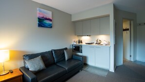 Una sala de estar moderna con un sofá de cuero negro, una mesita con una lámpara y un cuadro en la pared. Se observa una kitchenette con microondas, hervidor y pileta.