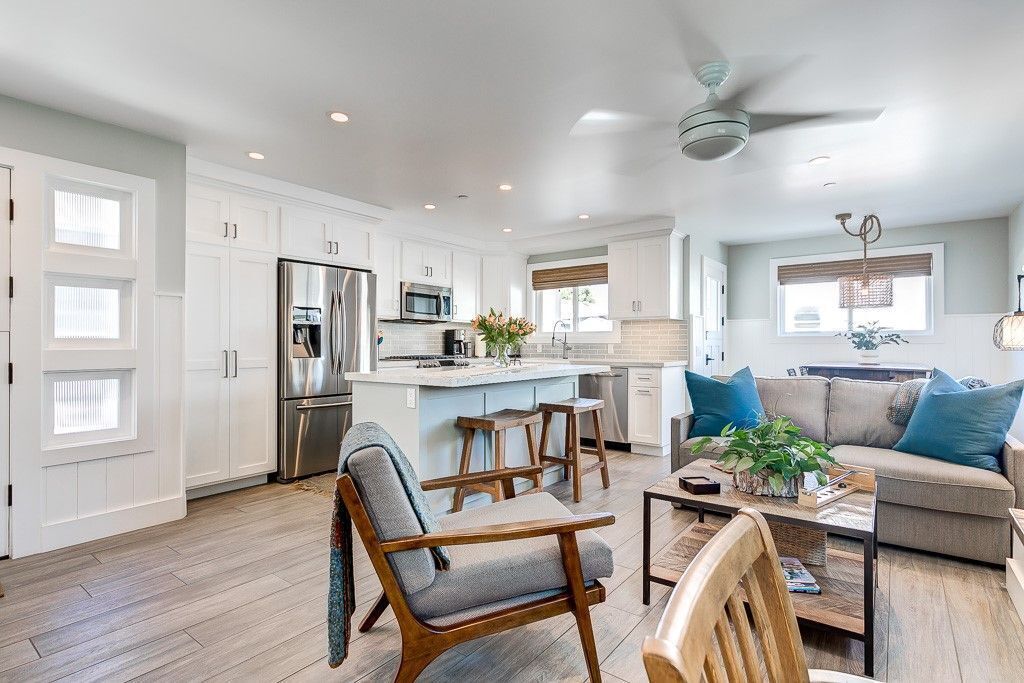 A spacious kitchen with white cabinets, stainless steel appliances, and a central island. There are wooden chairs and a sofa in the living area, with a ceiling fan above.