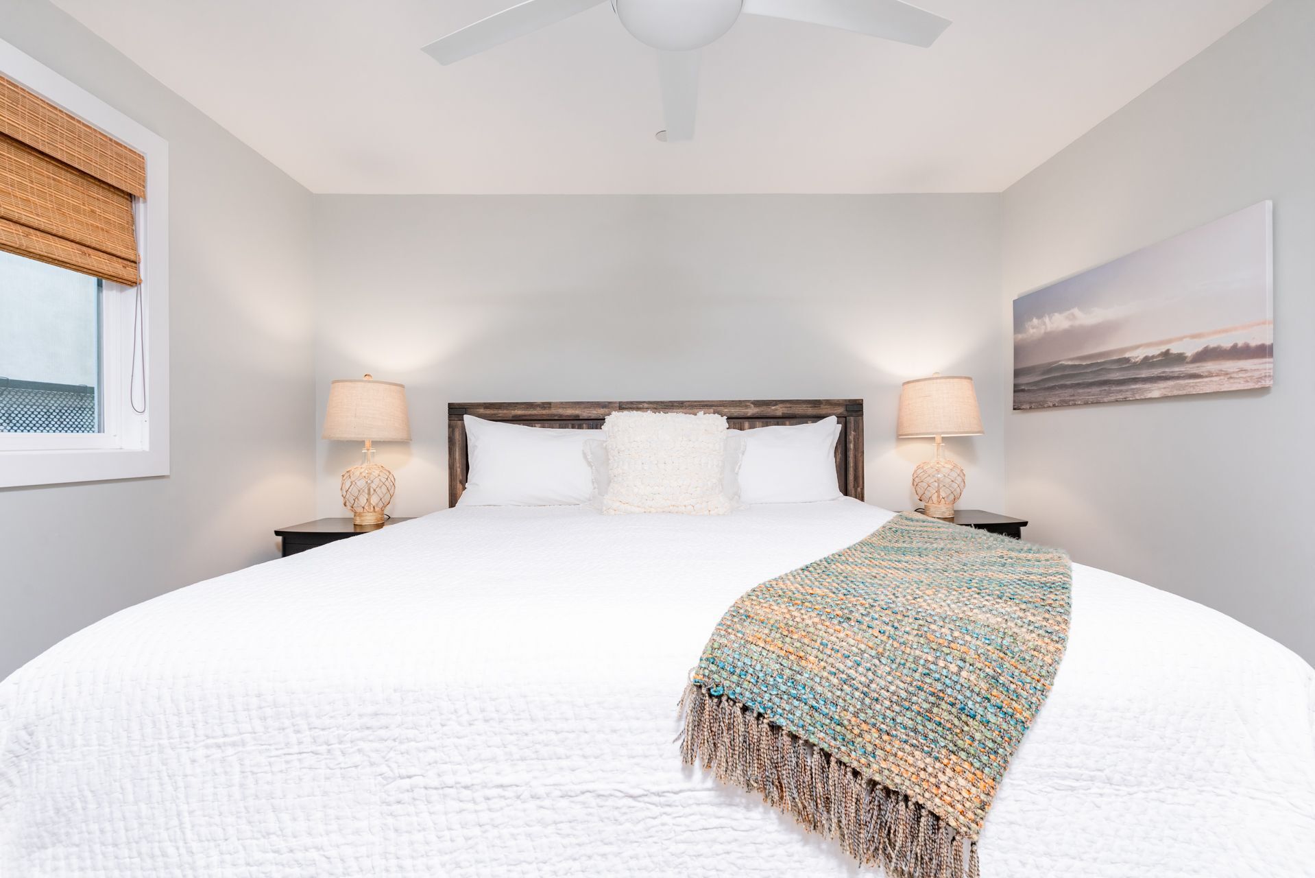 A bedroom with a large bed, two bedside tables with lamps, a ceiling fan, a window with a view, and a painting on the wall.