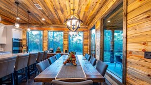 A wooden cabin interior with a dining table, chairs, a chandelier, and large windows. - Tranquil Haven - Sleeps 21 - Modern Rustic Luxury Retreat Among the Pines! (Hochatown)