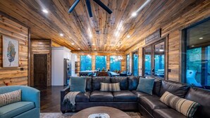 A modern cabin interior with a wooden ceiling, a leather sofa, a coffee table, and large windows offering a view of the outdoors. - Tranquil Haven - Sleeps 21 - Modern Rustic Luxury Retreat Among the Pines! (Hochatown)