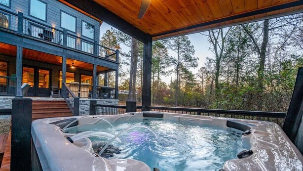 A hot tub on a wooden deck with a view of a modern building and trees. - Tranquil Haven - Sleeps 21 - Modern Rustic Luxury Retreat Among the Pines! (Hochatown)