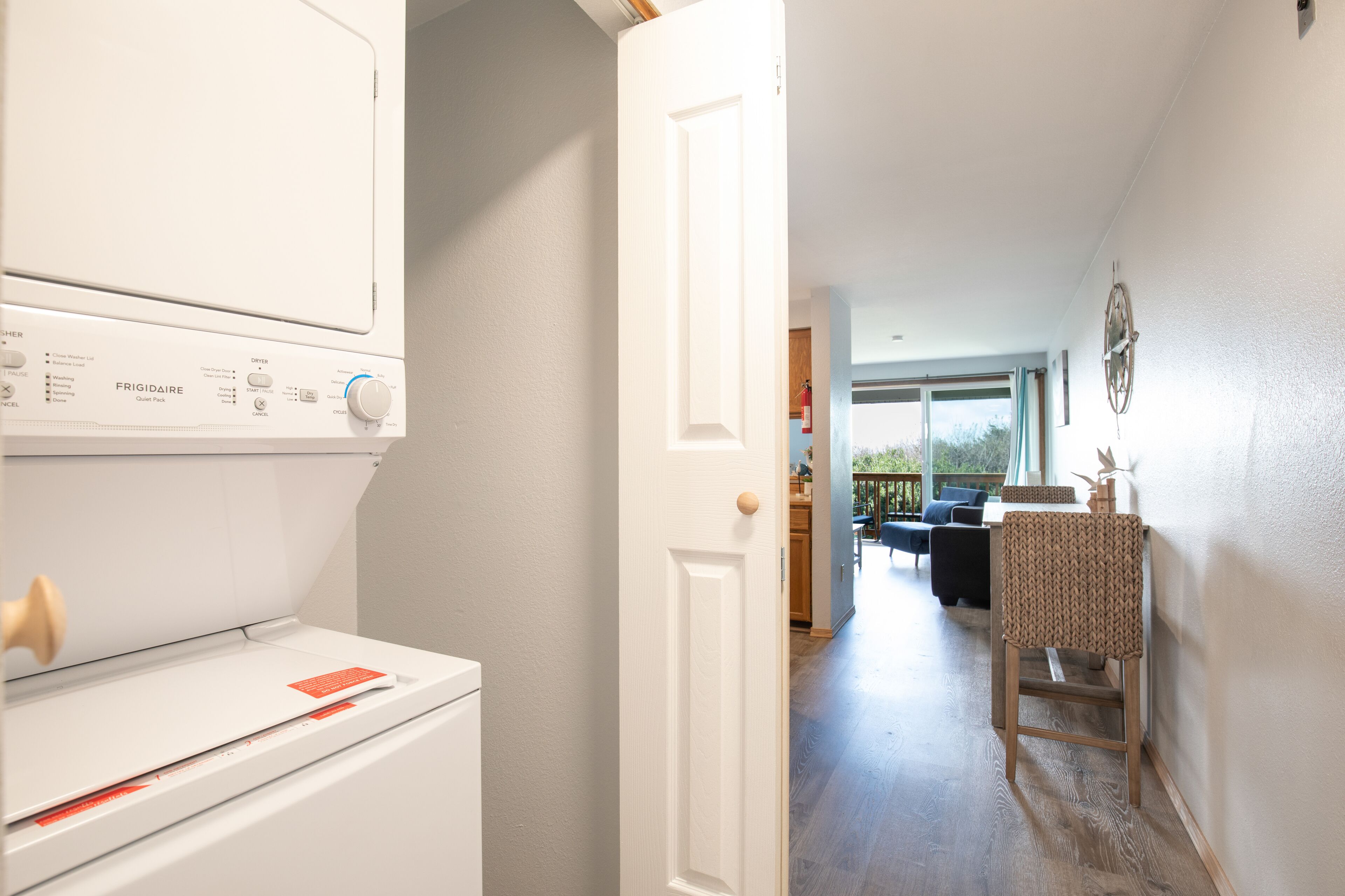 A laundry area with a front-loading washer and dryer, a wooden door leading to a living space with a view of the outdoors.