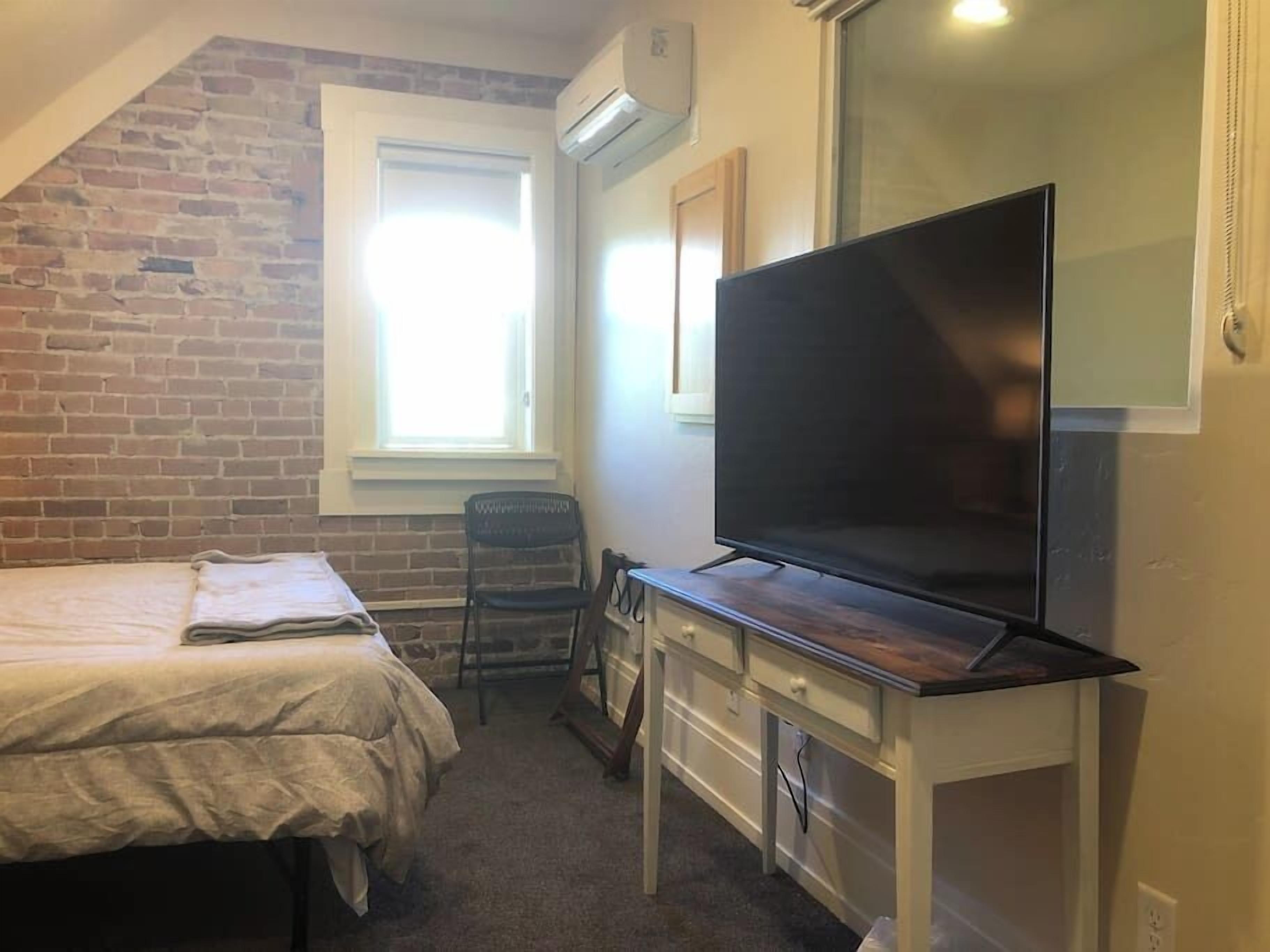 Une petite pièce comprenant un lit, un bureau avec un téléviseur à écran plat, une chaise et une fenêtre avec des stores.