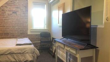 Une petite pièce comprenant un lit, un bureau avec un téléviseur à écran plat, une chaise et une fenêtre avec des stores.