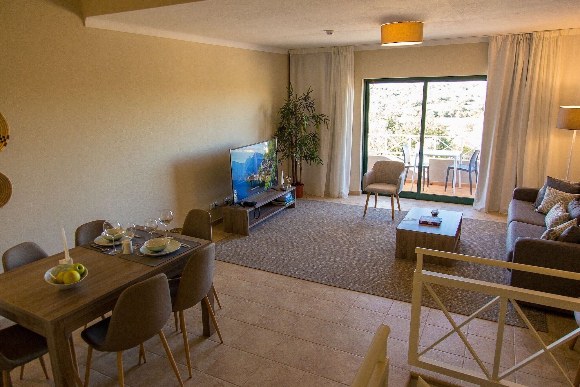 A modern living room with a dining area, a flat-screen TV, a potted plant, and a view of the outdoors.