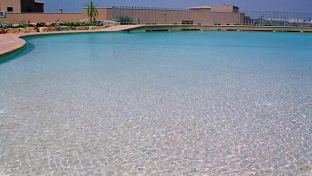 A clear pool with a curved edge, surrounded by a low wall and a fence. - Noria Velha (Lagoa)