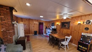 A wooden-paneled room with a dining table, chairs, a couch, a refrigerator, a stove, and a Pac-Man arcade machine. - Blakley Hobbit Large Cabin (Viola)