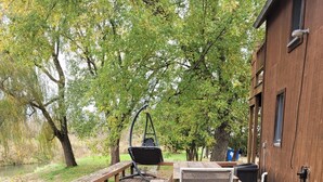A wooden deck with picnic tables and a hanging chair, surrounded by trees and a building. - Blakley Hobbit Large Cabin (Viola)