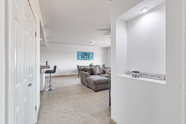 A modern living room with a grey sofa, a dining area with a bar stool, and a kitchen area with a marble countertop.