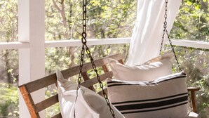 Una silla mecedora de madera con cojines y una manta de macramé, suspendida en un porche con vistas a árboles.