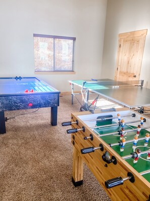 Una habitación con dos mesas de fútbol de mesa y una mesa de ping pong, suelo alfombrado, puerta de madera y una ventana con persianas.