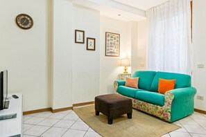 A living room with a teal sofa, a brown ottoman, a television, and framed pictures on the wall.