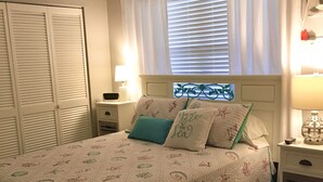 Ein Schlafzimmer mit Bett, Vorhängen, einem Fenster mit Jalousien, einem Nachttisch und einem Kleiderschrank.