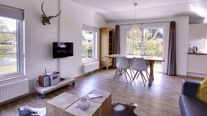 Un salon moderne avec un téléviseur, une table basse en bois, un coin repas avec des chaises et une cuisine en arrière-plan.