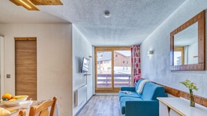 Ein modernes Wohnzimmer mit einem blauen Sofa, einem Holzespeisetisch und Stühlen. Ein Fernseher ist an der Wand montiert, ein großes Fenster bietet Blick auf Gebäude, und die Decke besteht aus Holz.