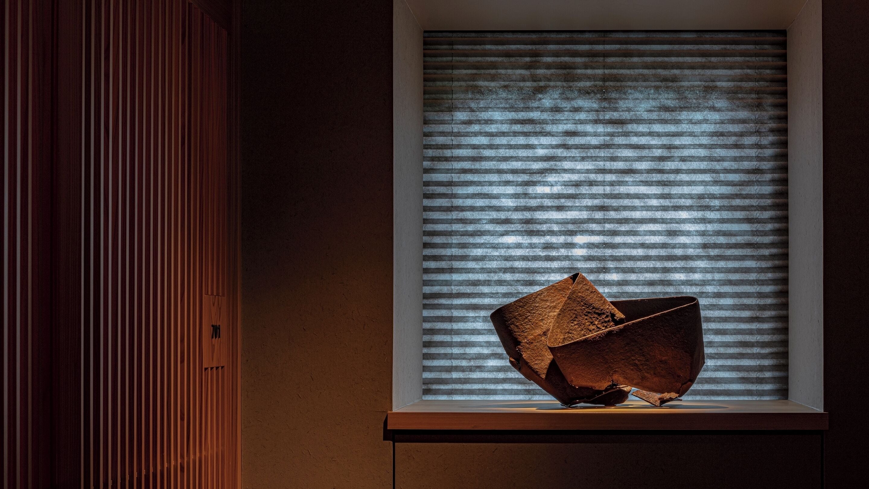 A room with a window featuring horizontal blinds, a wooden cabinet, and a sculpture on a pedestal.