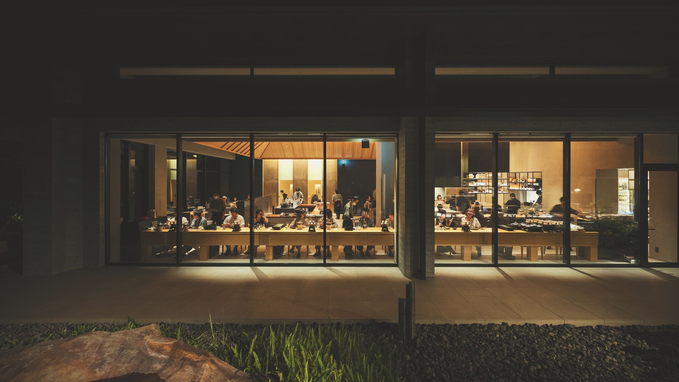 A modern building with large glass windows, a spacious interior with tables and chairs, and a visible kitchen area.