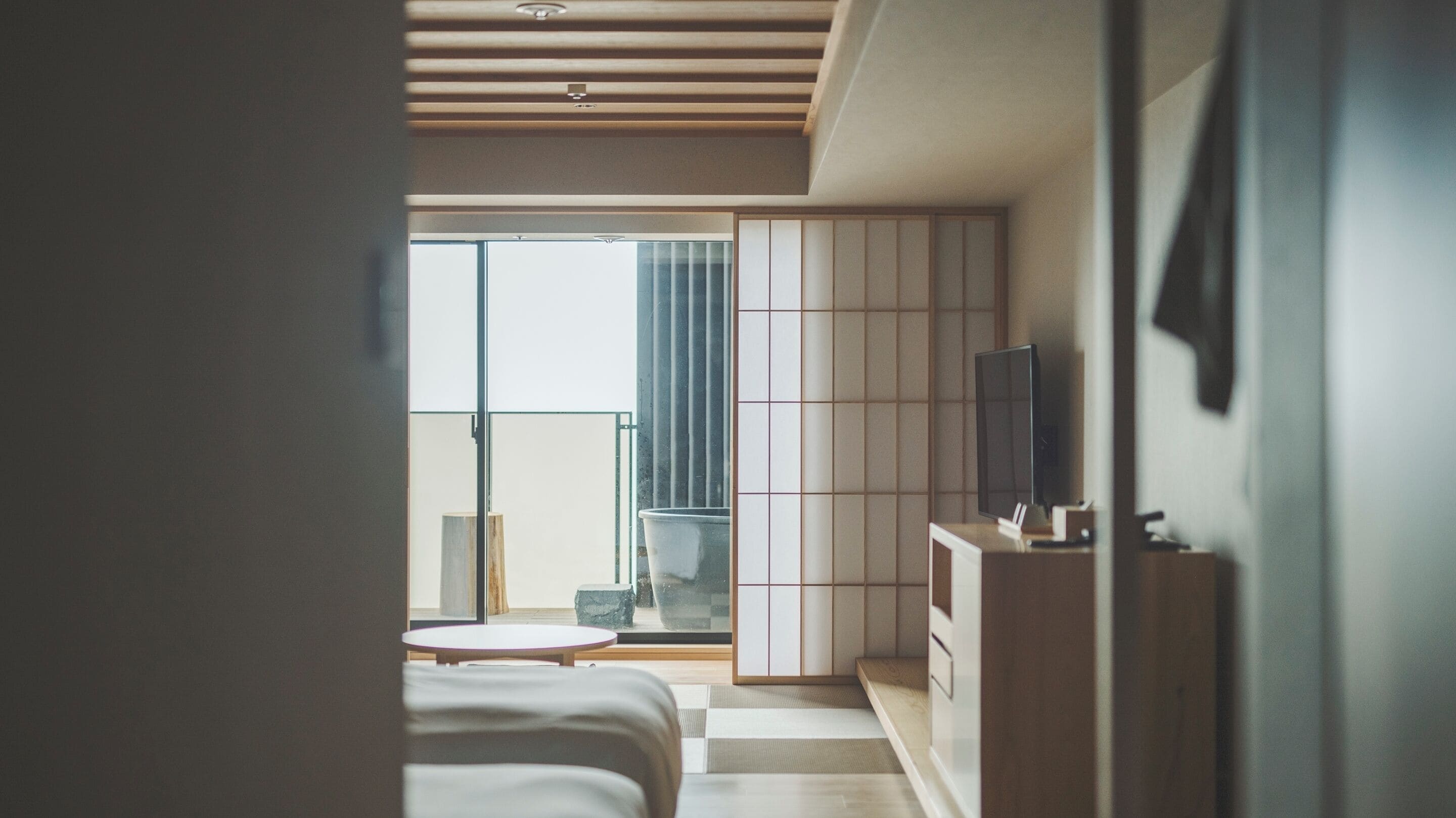 Ein modernes Hotelzimmer mit einer großen Schiebeglasdoor zum Balkon, einem Flachbildfernseher, einem kleinen runden Tisch und einem traditionellen japanischen Kleiderschrank.