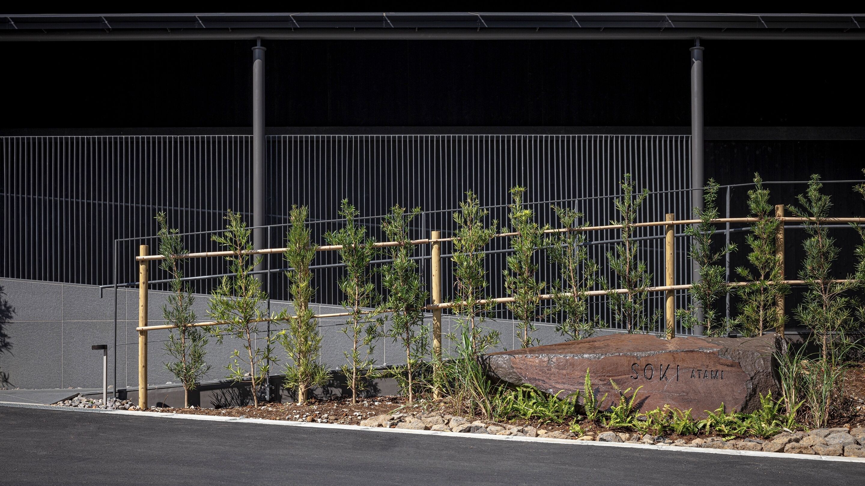 A landscaped area with young trees, a large rock with 'SOKIMAMI' engraved, and a metal fence.