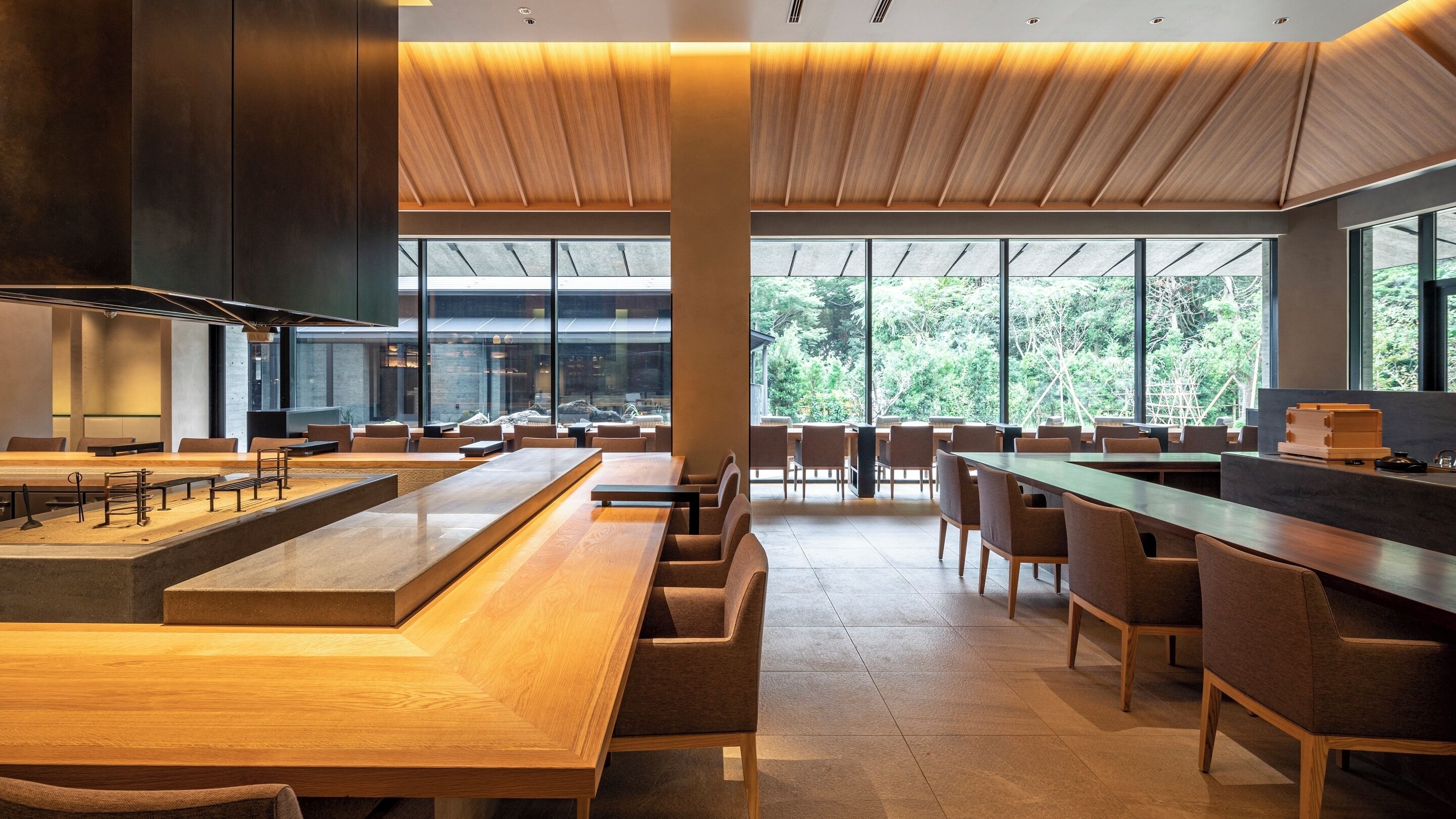 A modern restaurant with wooden tables and chairs, large windows, and a view of greenery.
