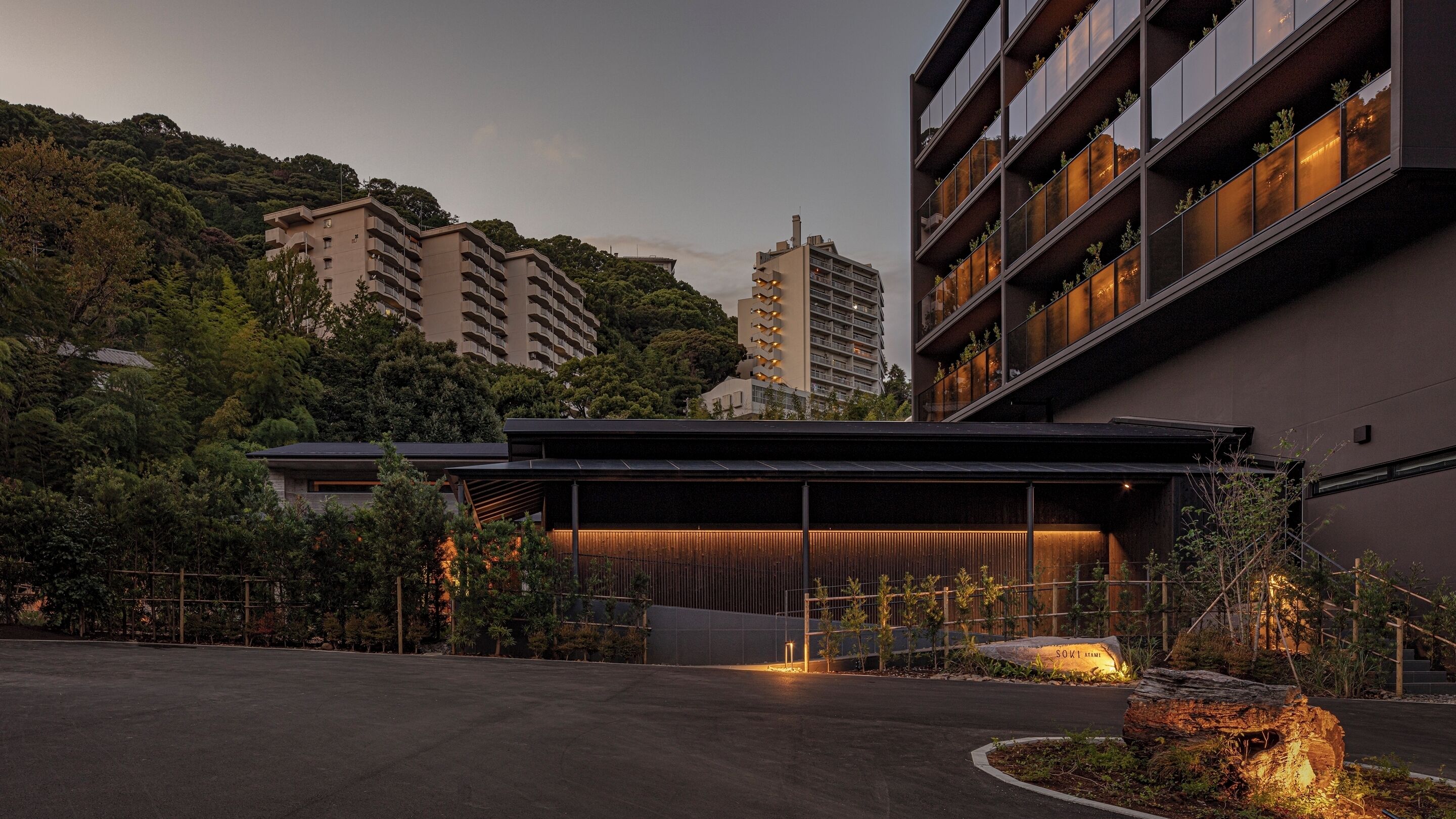 A modern hotel entrance with a covered outdoor area, surrounded by lush greenery and adjacent high-rise buildings.