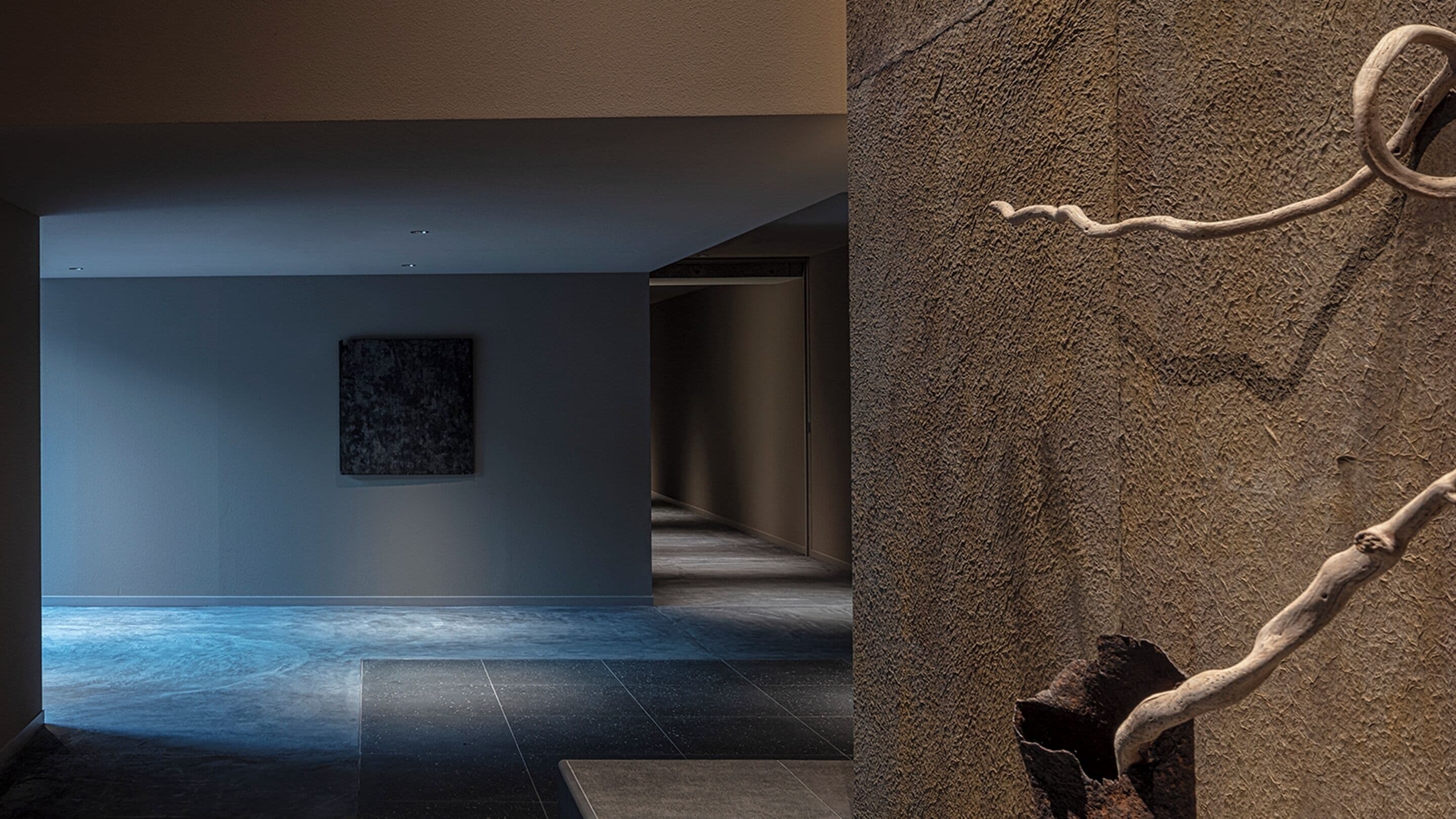 A hallway with a dark floor, a framed artwork on the wall, and a textured wall with a carved branch.