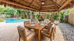 A covered outdoor dining area with a pool, wooden furniture, and a thatched roof. - Hidden Gem Villa | Heated Pool, Hot Tub & Tikihut (Fort Lauderdale)