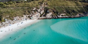 An aerial view of a beach with clear turquoise waters, rocky cliffs, and a road running parallel to the beach.