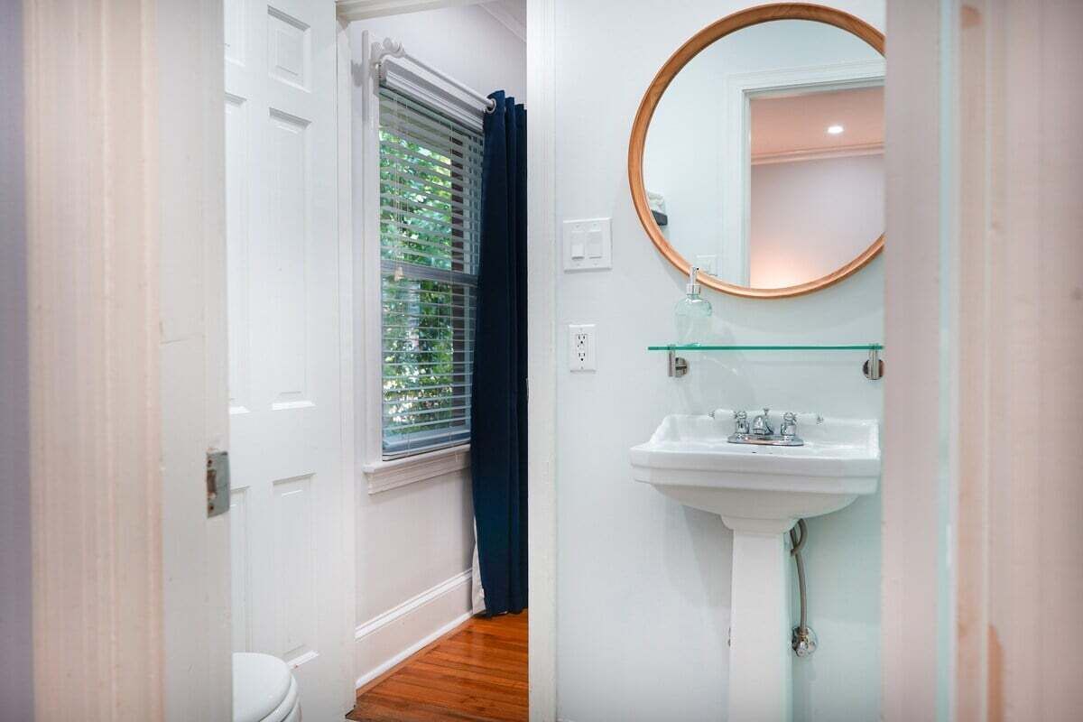 A bathroom with a white sink, a round mirror, and a window with blinds.