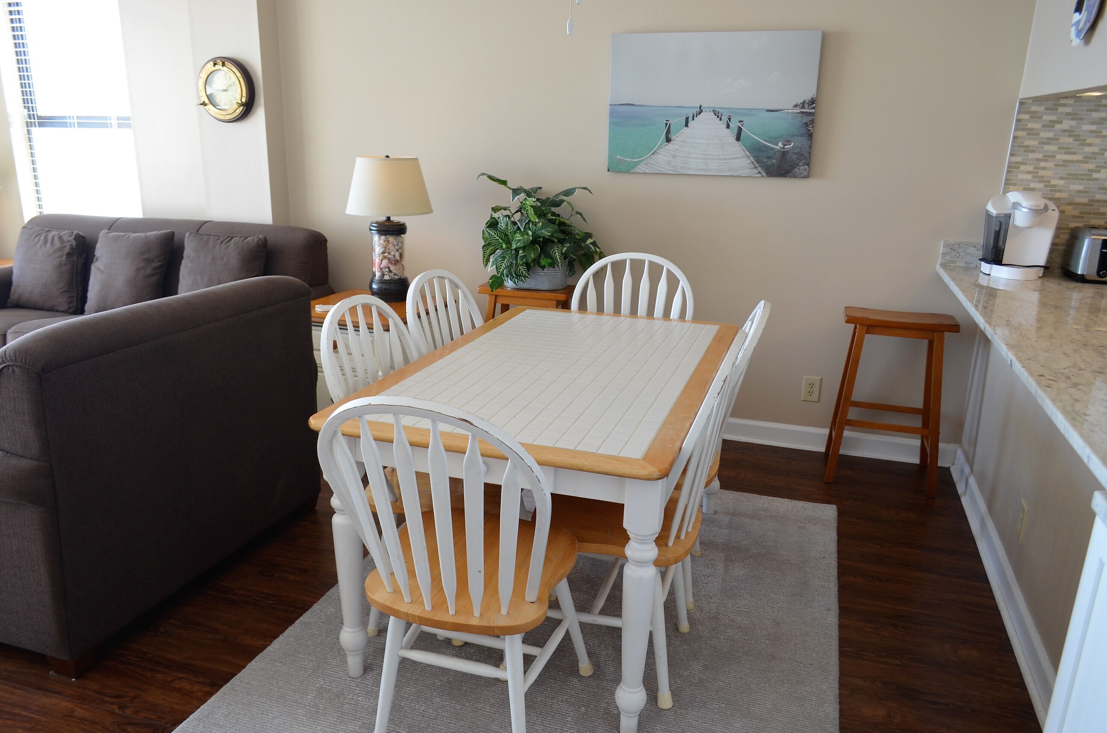A dining area with a wooden table and white chairs, a grey rug, a lamp, a potted plant, and a painting on the wall.