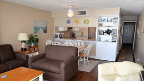 Ein Wohnzimmer mit braunem Sofa, einem weißen Sessel und einem Holztisch. Es gibt eine Küchenzeile mit weißen Schränken und einem Kühlschrank.
