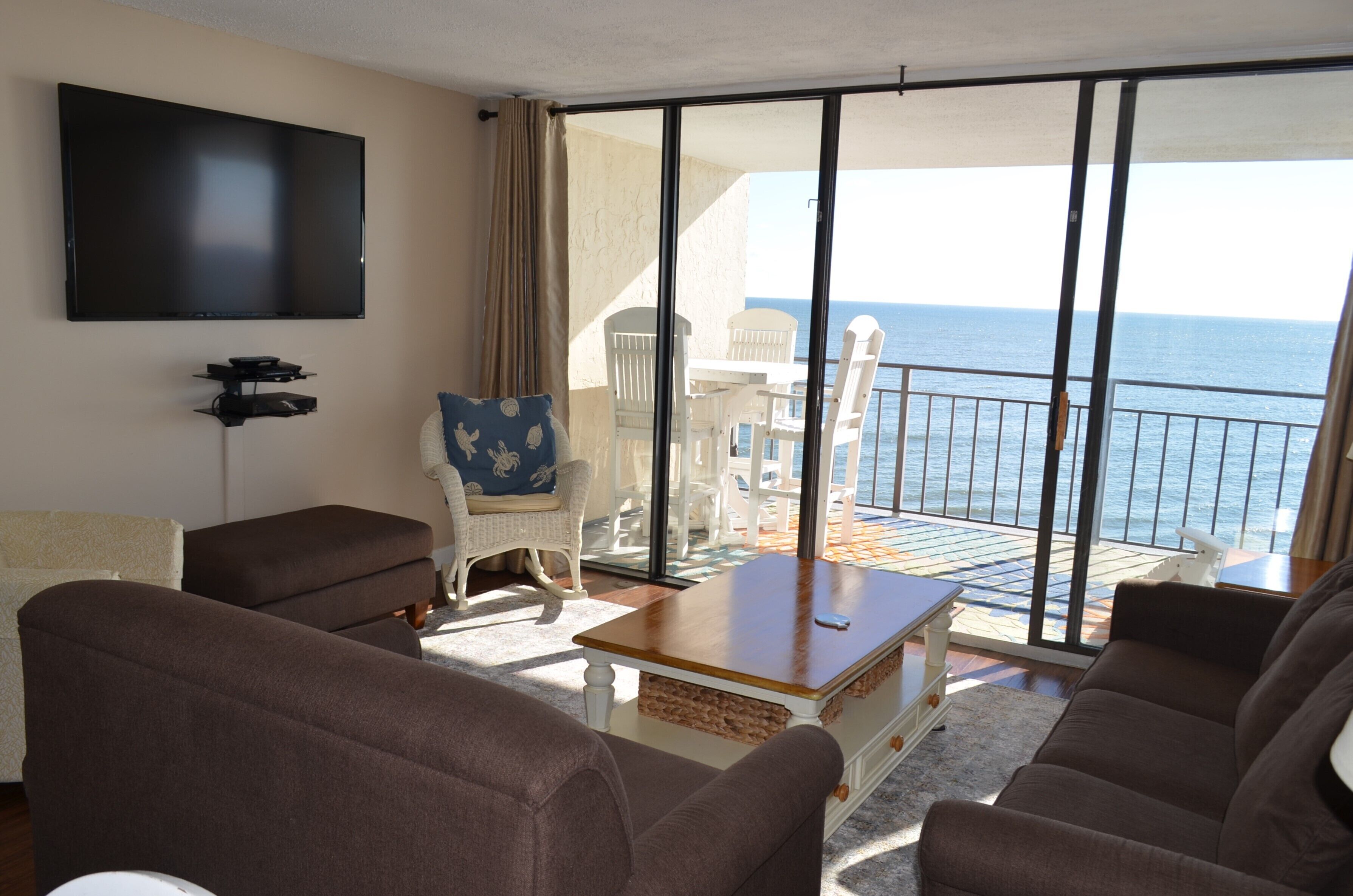 A living room with a large TV, comfortable seating, and a balcony with a view of the sea.