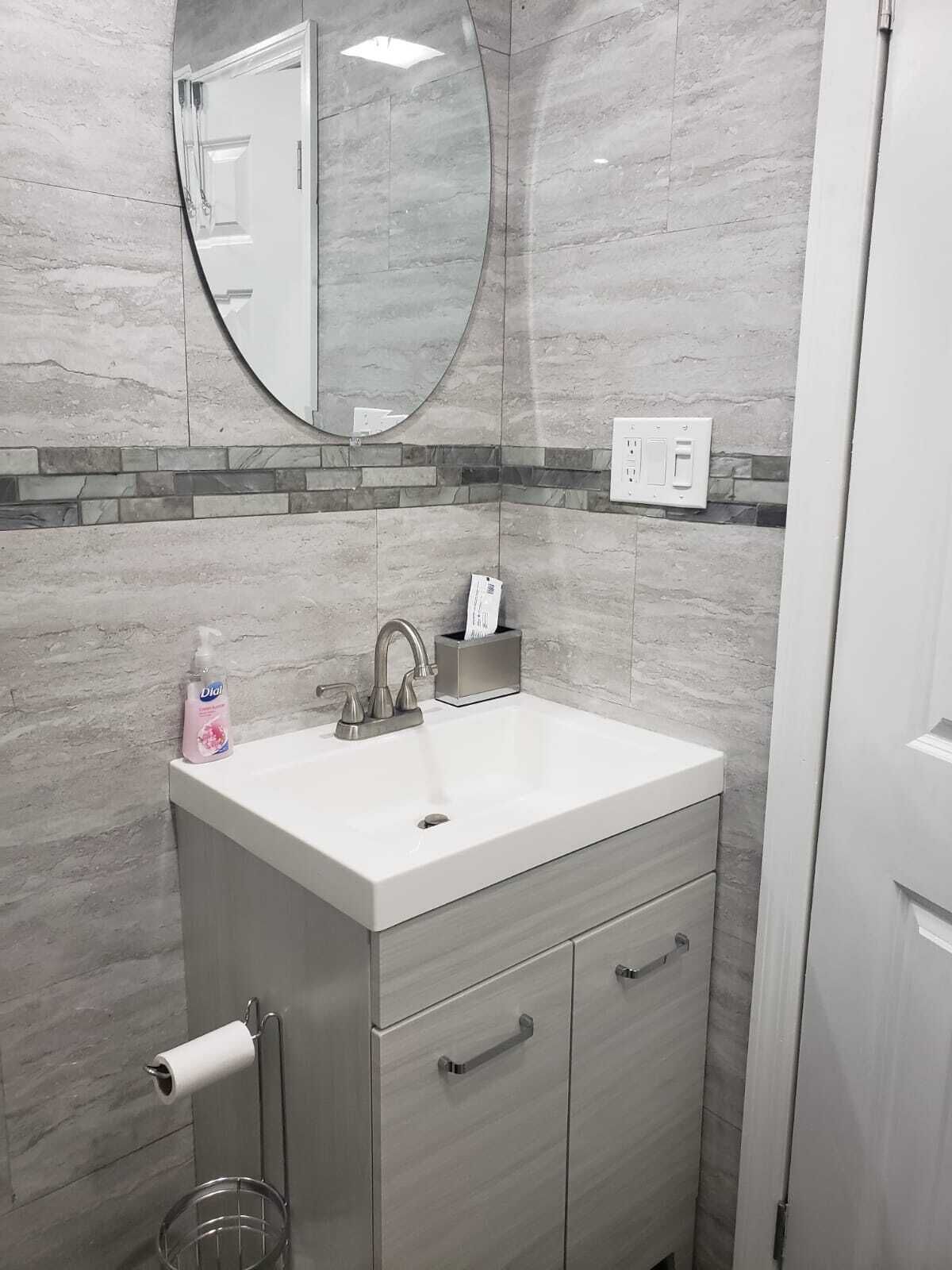 Un baño con lavamanos blanco, un espejo, dispensador de papel higiénico y un cubo de basura.