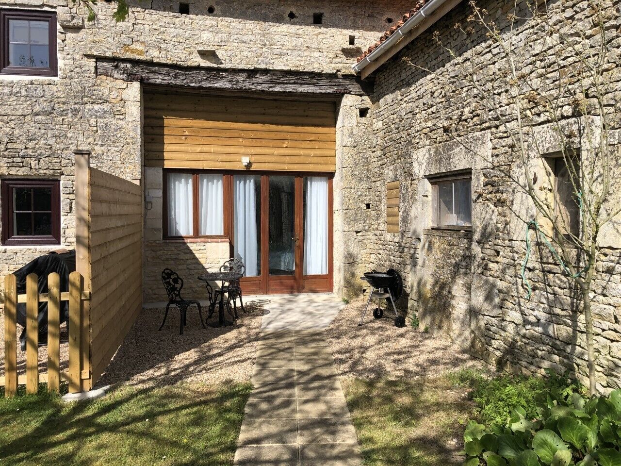 A stone building with a wooden door and windows, a patio with a table and chairs, and a barbecue grill.