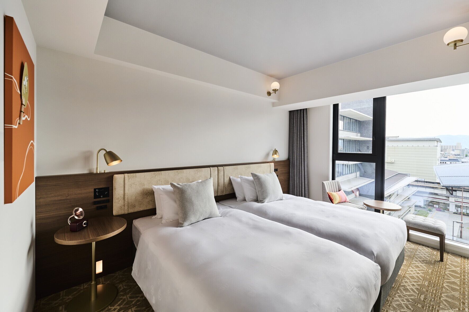 A hotel room with two beds, a small table, a chair, a desk, a lamp, and a large window with a view.