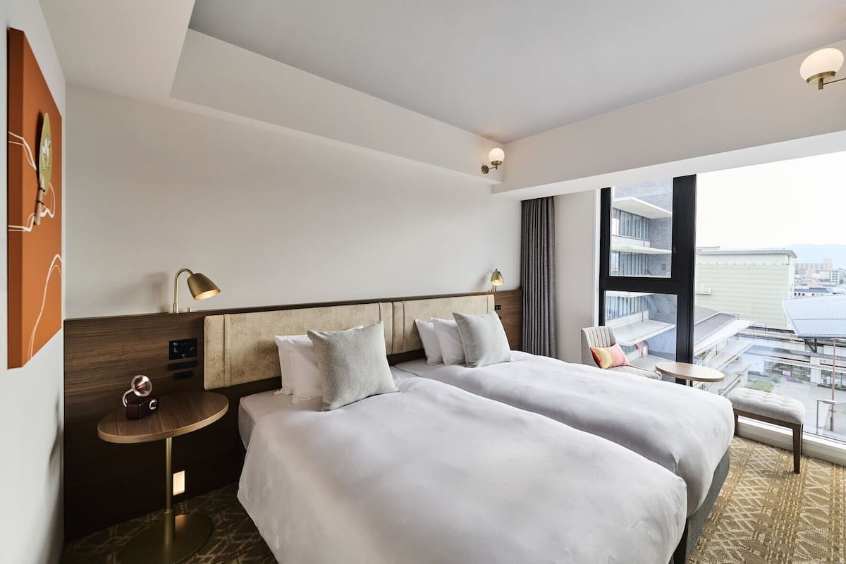 A hotel room with two beds, a small table, a chair, a desk, a lamp, and a large window with a view.