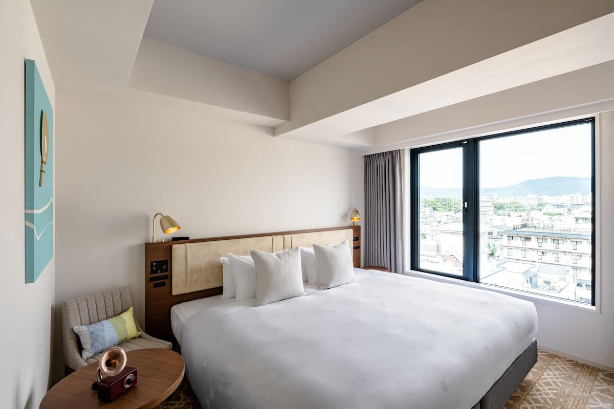 A hotel room with a large bed, a nightstand with a lamp, a chair with a cushion, and a view of the city through large windows.