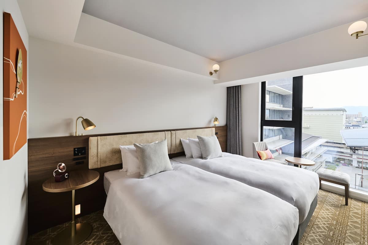 A hotel room with two beds, a small round table, a lamp, and a view of the city through large windows.