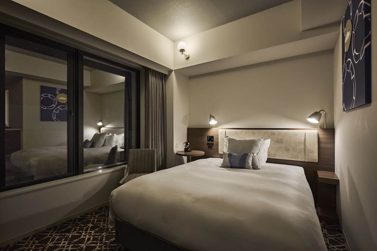 A hotel room with a large bed, bedside lamps, a nightstand, a window with curtains, and a wall art piece.
